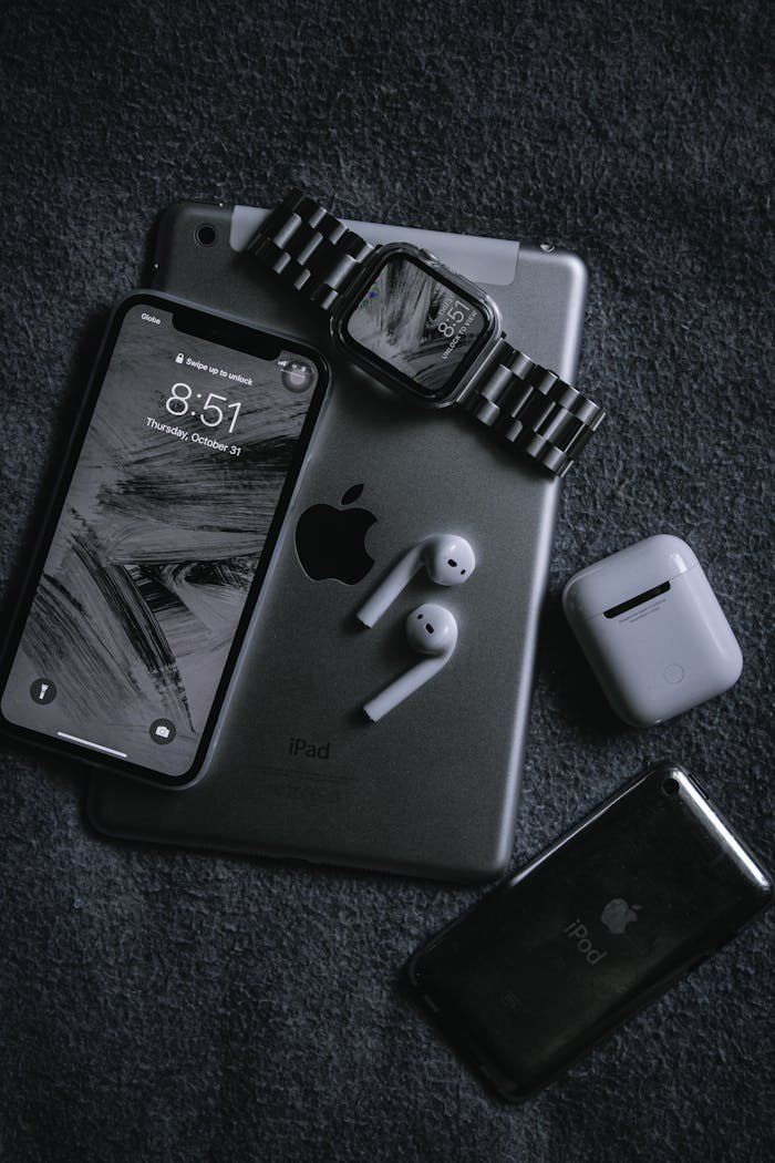 Black and white tech gadgets arranged artistically, including a phone, earbuds, tablet, and wristwatch.