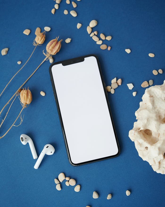 Flat lay of smartphone with wireless earphones on blue surface with natural elements.